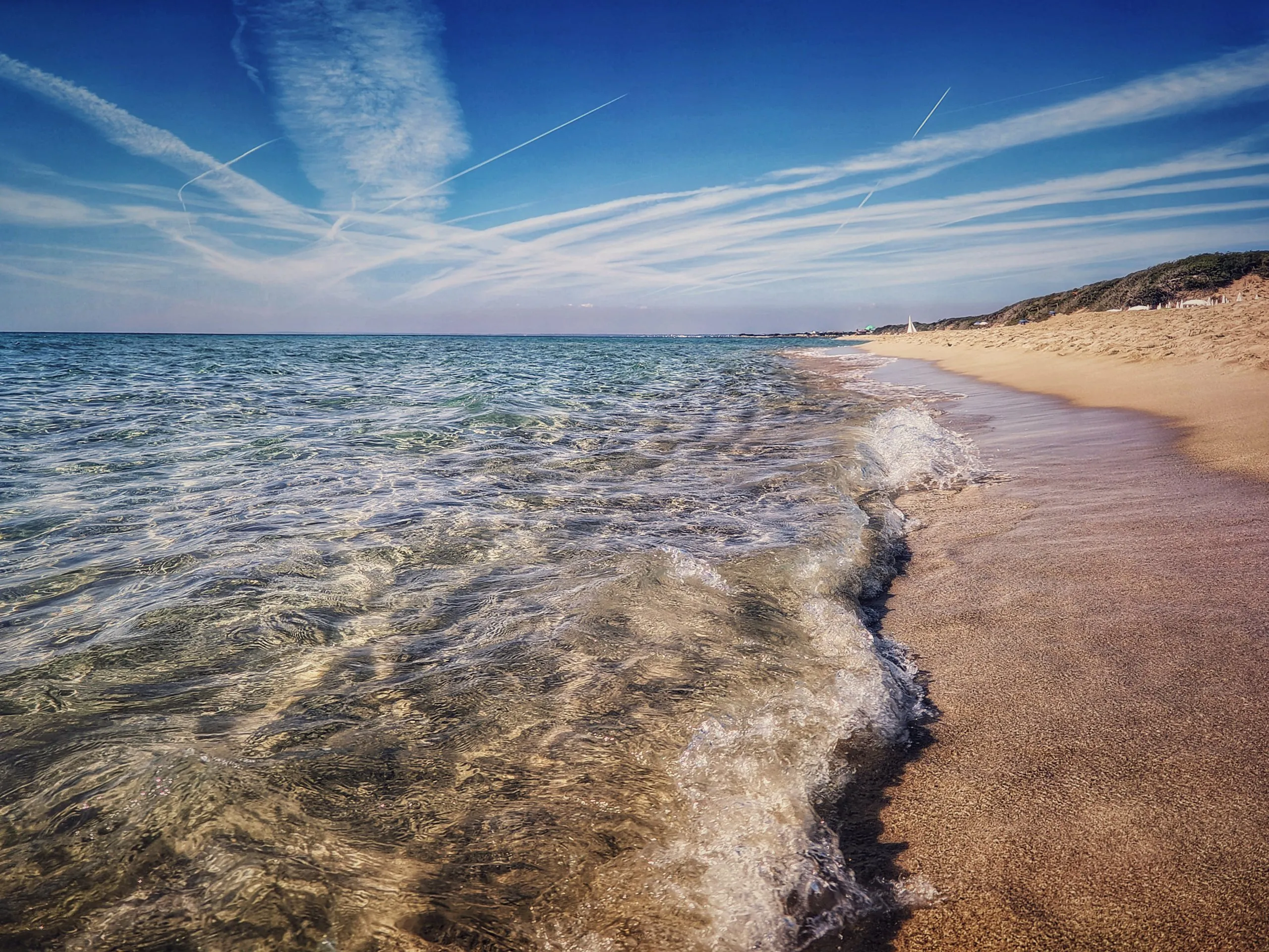La battigia con le onde che si stendono sulla sabbia bagnata, le dune con la macchia bassa sulla destra e il cielo striato