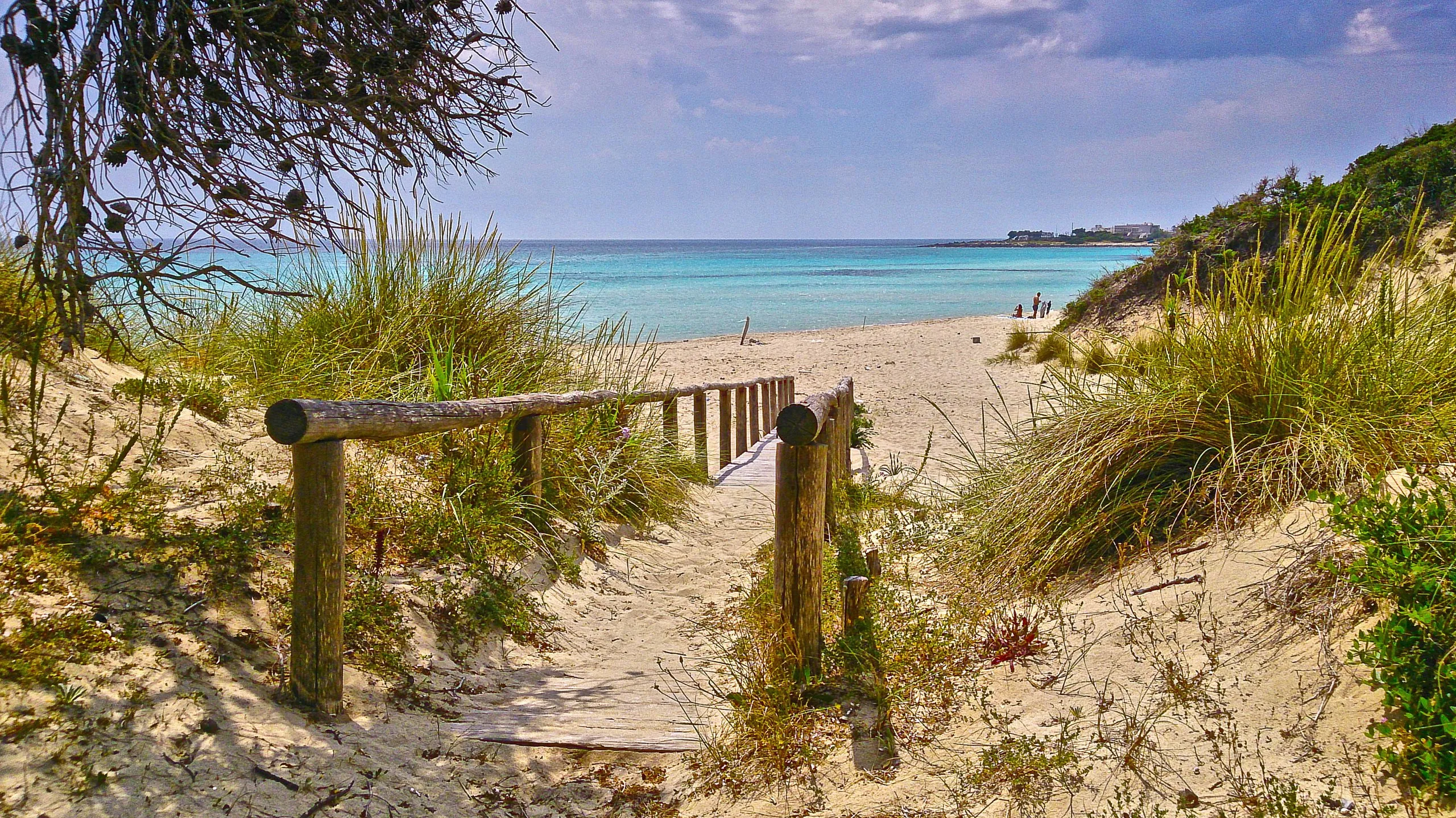 Una passerella di legno con la staccionata fra le dune coperte di macchia, e in fondo la spiaggia e il mare turchese