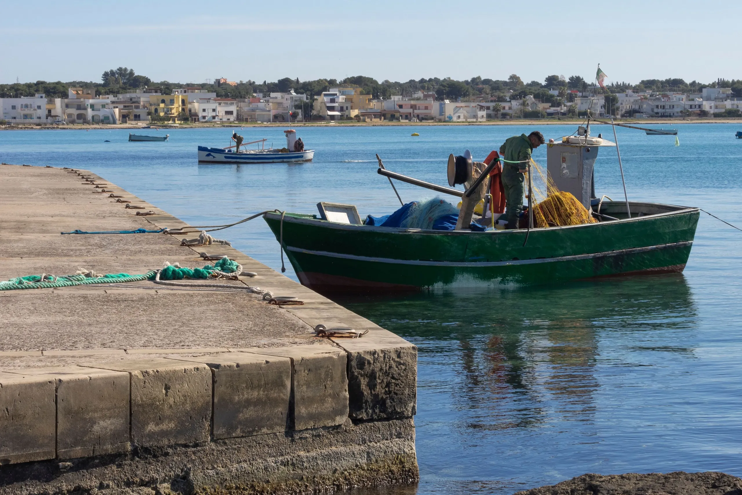 Un pescatore al lavoro sulla sua barca ormeggiata al molo, con il paese basso e bianco sull’altra sponda