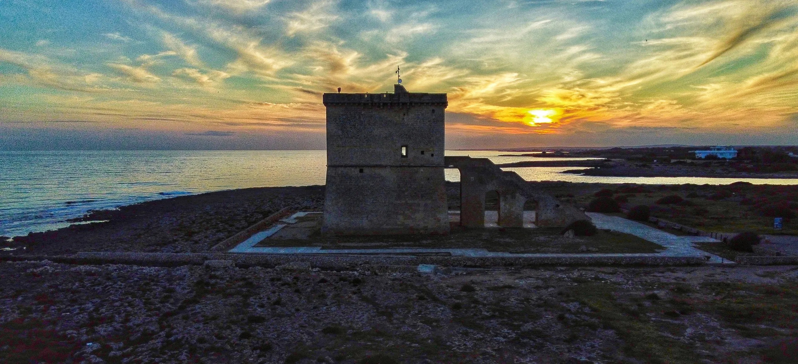 Torre Lapillo al tramonto: la torre quadrata di pietra sulla costa bassa, il sole all’orizzonte e il mare sulla sinistra