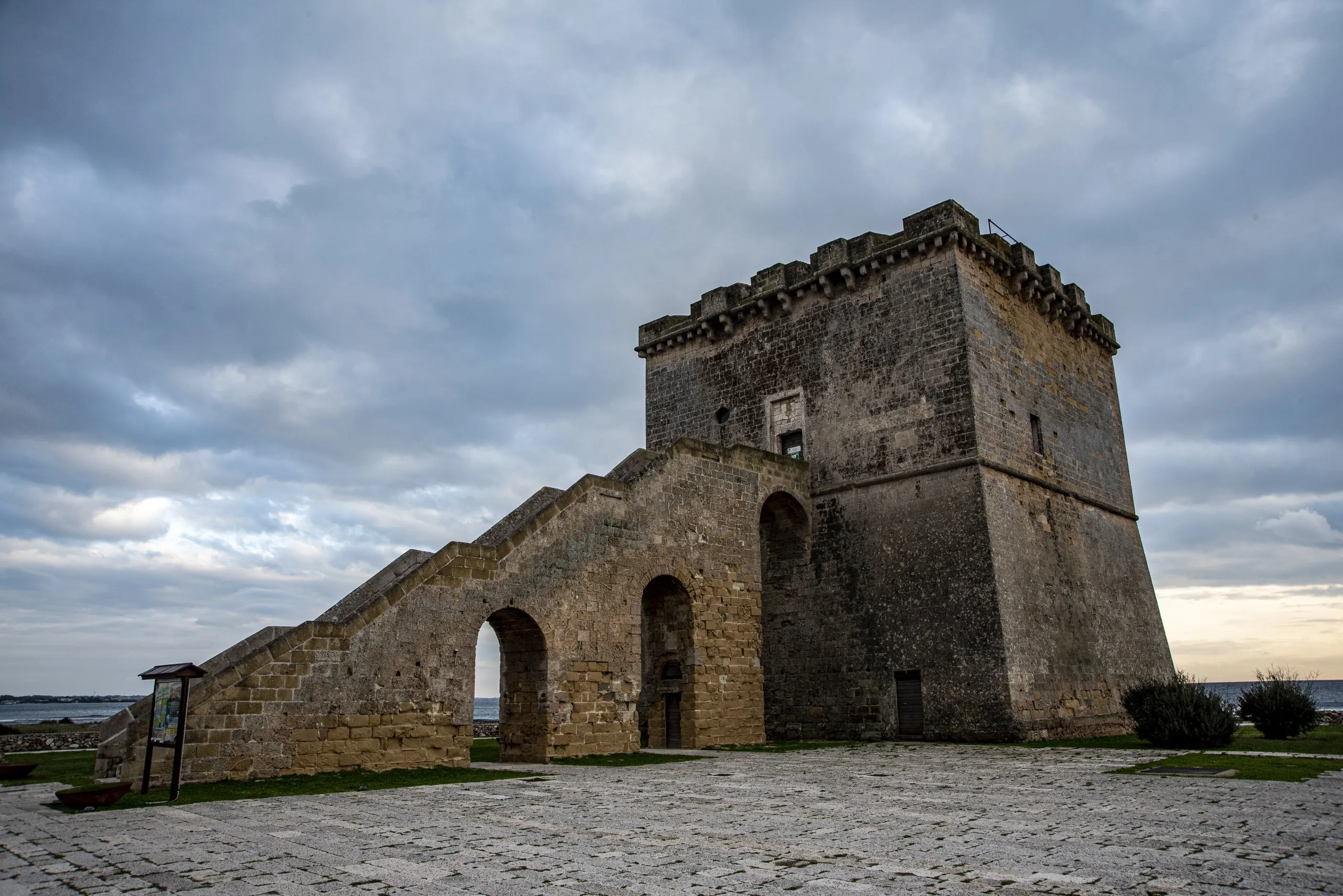 Una torre costiera quadrata di pietra, con la rampa ad arcate che sale fino al portale e il cielo nuvoloso sopra